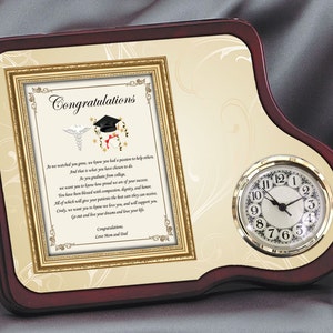 May include: A wooden plaque with a gold frame and a clock on the right side. The plaque has a cream background with a gold design and a poem inside the frame that reads "Congratulations. As we watched you grow, we knew you had a passion to help others. And that is what you have chosen to do. As you graduate from college, we want you to know how proud we are of your success. You have been blessed with compassion, dignity, and honor. All of which will give your patients the best care they can receive. You have been blessed with compassion, dignity, and honor. All of which will give your patients the best care they can receive. You will have your patients' love, and they will support you. Only, we want you to know we love you, and will support you. Go out and live your dreams and love your life. Congratulations. Love Mom and Dad."