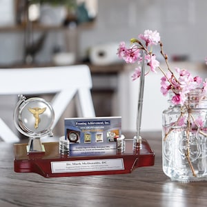 May include: A wooden desk organizer with a silver globe featuring a medical symbol, a pen holder, and a business card holder. The organizer has a plaque with the text "Dr. Mark McDonalds, DC" and the text "Framing Achievement, Inc." on a business card. A mason jar with pink flowers sits on a wooden table.