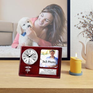 May include: A mahogany wood clock and photo frame with a silver clock face and a 3x3 photo space. The clock face has a silver caduceus symbol and the text "Clock & Thomas, OD". The frame has a silver inscription that reads "Enjoy a monthly reminder for one year of service at the New York Eye and Ear Infirmary.  Clock & Thomas, OD.  June 2018".