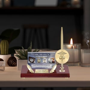 May include: A mahogany desk set with a gold pen and a gold circular emblem with a caduceus symbol. The emblem is engraved with the letters "O.D." The desk set is on a wooden table with a business card that reads "Framing Achievement, Inc. www.ARGiftFrames.com Premium Engraved Clocks, Retirement Gifts, Competition Service Awards and Personalized Poetry Frames Dr. Jaime Walker, Ohio Engraved Clocks by ARGiftFrames.com".