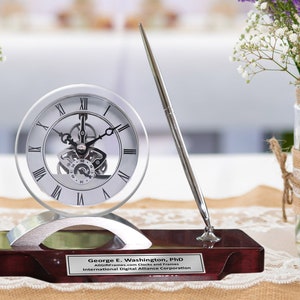 May include: A silver desk clock with a visible gear mechanism and a silver pen stand on a mahogany base with a personalized plaque that reads "George E. Washington, PhD AllGiftFrames.com Clocks and Frames International Digital Alliance Corporation".