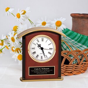 May include: A mahogany desk clock with a gold trim and a white face. The clock has Roman numerals and a gold-colored second hand. The clock is on a gold base and has a plaque with the text "Cindy K. McDaniels Engraved Desk Clocks by AllGiftFrames.com Distributor of Recognition Clocks and Personalized Gifts".