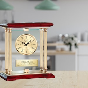 May include: A gold-toned desk clock with Roman numeral markings on a gold face. The clock is encased in a glass frame with gold pillars and a mahogany base. The inscription reads "Kylie C. Lee, CPA, Certified Public Accountant, Los Angeles Accounting Services Corp, Engraved Desk Clocks by AllGiftFrames.com".