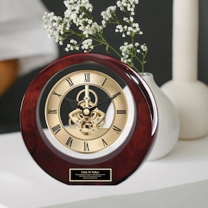 May include: A round, wooden desk clock with a transparent face, showcasing the inner workings of the clock. The clock has a gold-colored face with Roman numerals and a dark red wooden frame. A small plaque at the bottom has text. A vase with white flowers is in the background.