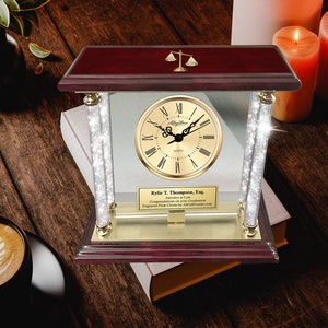 May include: A mahogany desk clock with a gold-tone face and a glass front. The clock has a scale of justice on top and a gold-tone base with a personalized inscription. The inscription reads "Rylie T. Thompson, Esq. Attorney at Law Congratulations on your Graduation Engraved Desk Clocks by AllGFrames.com".