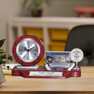 May include: A mahogany desk clock with a silver face and black hands. The clock is on a mahogany base with a silver plaque that reads "Dr. Jadyn C. Smith, OD Doctor of Optometry ARGiftFrames.com Engraved Clocks and Frames". There is a silver business card holder with a silver medical symbol on top. The business card holder has a business card that reads "Framing Achievement, Inc. www.ARGiftFrames.com".