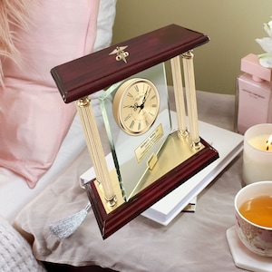 May include: A mahogany wood clock with gold accents and a glass face. The clock has a gold medical symbol on top and a personalized plaque with the name "Michael C. Walker, MD" on the bottom.