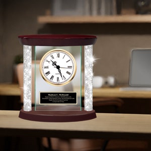 May include: A quartz desk clock with a gold-tone face, Roman numerals, and black hands. The clock is encased in clear glass with sparkling crystal accents and a dark wood base and top. A small plaque with text is visible.