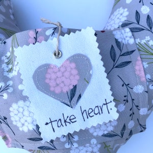 May include: A gray fabric heart-shaped tag with a pink floral design inside the heart. The tag has a white border and the words "take heart" written in black.