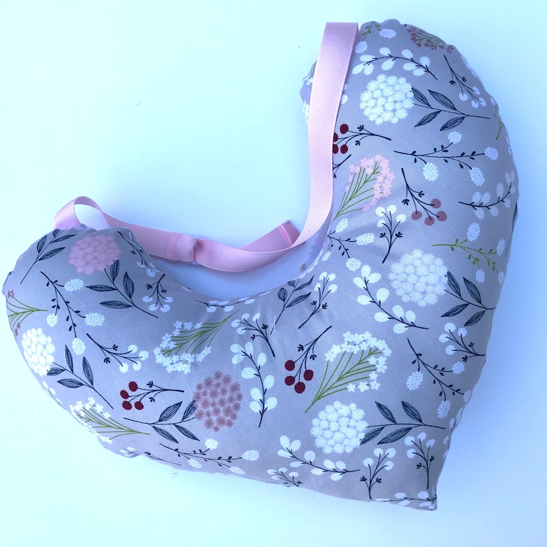 May include: A gray and white floral print pregnancy pillow with a pink strap. The pillow is shaped like a crescent moon and is designed to support the body during pregnancy.