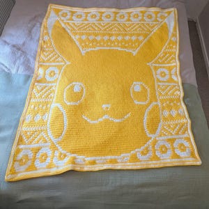 Puede incluir: Manta de ganchillo amarilla y blanca hecha a mano con un diseño de Pikachu. La manta tiene un borde detallado con patrones geométricos. La cara de Pikachu está en el centro, con sus características icónicas.