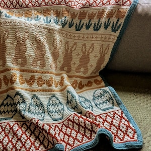 Pode incluir: Um cobertor de crochê creme com tema de Páscoa. O cobertor apresenta padrões repetidos de coelhos, ovos e desenhos florais em tons de marrom, laranja, verde-azulado e vermelho. A borda é um padrão de diamante com uma borda verde-azulado.