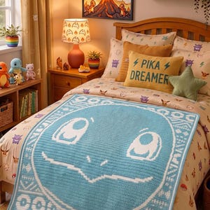 May include: A light blue crochet blanket featuring a large white outline of a character face with a geometric border, draped over a bed with patterned bedding, a yellow pillow with the text PIKA DREAMER, and various character-themed decor.