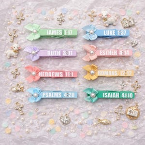 May include: A collection of colorful wooden clothespins, each adorned with a satin bow and a pearl. The pins feature Bible verses like James 1:17 and Isaiah 41:10. Decorative crosses and a lamb figurine are scattered around.