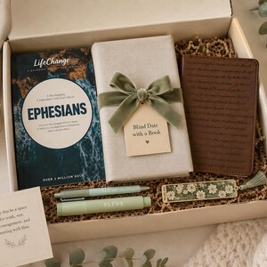 May include: A gift box containing a LifeChange Ephesians book, a book wrapped in beige paper with a sage green velvet bow and a tag reading Blind Date with a Book, a brown journal, two sage green pens, a floral bookmark, and a card with printed text.