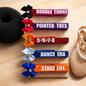 May include: A collection of decorative clothespins with text and bows, including "DOUBLE TURNS," "POINTED TOES," "5•6•7•8," "DANCE ERA," and "STAGE LIFE." Each pin has a different color and bow. A black hair donut and a ballet shoe are also in the image.