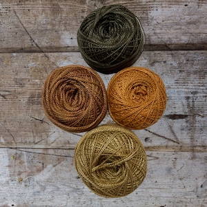 May include: Four balls of yarn in different shades of brown and green. The yarn is arranged in a circle on a wooden surface.