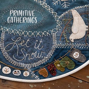 May include: A blue and white felt Christmas tree skirt with the text "Let it Snow" and a white dove with a brown branch. The skirt is decorated with white snowmen, brown mittens, and green and red mittens.