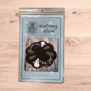 May include: A blue and white pattern with the text "Nutmeg Hare" and "#9 Dress me up Frosty". The pattern features a circular design with a snowman, trees, and other winter-themed elements.