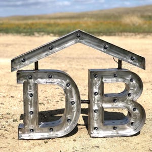 May include: Metal letters spelling "DB" with a metal roof-shaped structure above them. The letters have small round holes throughout, giving the appearance of a light-up sign.