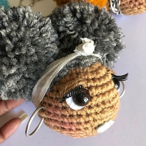 May include: Crocheted keychain with a brown face, white eyes, black eyelashes, and a silver headband. The keychain has gray pom-poms for hair.
