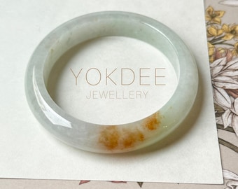 May include: A jade bangle bracelet with a pale green and orange hue. The bracelet has a smooth, polished surface and is circular in shape. The text "YOKDEE JEWELLERY" is printed on the surface.