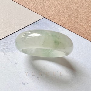 May include: A translucent jade ring with a soft green hue. The ring is smooth and polished, showcasing the natural variations in the stone. The ring is set against a neutral background.
