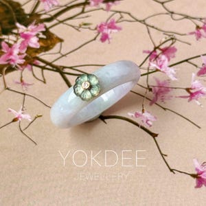 以下が含まれることがあります： 淡い緑色の翡翠バングルブレスレット。花型のアクセントが付いています。花は緑色の花びらのデザインで、中央に小さく丸い透明な石があります。ブレスレットは、ピンクの花と枝を背景に配置されています。