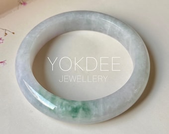 Puede incluir: Un brazalete de jade liso y circular con una base gris claro translúcida y una mancha distintiva de color verde vibrante. El texto YOKDEE JEWELLERY está impreso en blanco en el centro del brazalete, el cual tiene un acabado pulido.