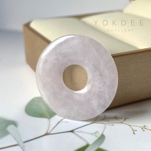Könnte beinhalten: Ein blass-lavendelfarbener Jade-Donut-Anhänger mit einer glatten, polierten Oberfläche. Der Anhänger wird auf einer beigefarbenen Samtbox mit dem aufgedruckten Text "YOKDEE JEWELLERY" präsentiert.