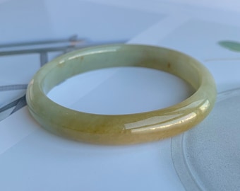 May include: A smooth, polished bangle bracelet crafted from translucent jade stone. The circular accessory features a gradient of pale green and warm yellow hues, with a glossy, reflective surface that highlights the natural mineral variations.