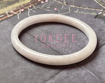 May include: A smooth, polished, off-white jade bangle bracelet with a rounded profile. The surface of the bangle has a subtle, cloudy texture. The text YOKDEE JEWELLERY is overlaid in pink lettering across the center of the circular bangle.