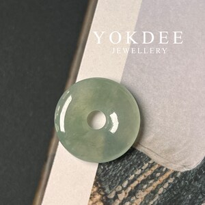 Könnte beinhalten: Ein hellgrüner Jade-Anhänger in Form eines Donuts mit einem Loch in der Mitte. Der Anhänger liegt auf einer weißen Oberfläche mit dem aufgedruckten Text "YOKDEE JEWELLERY".