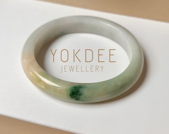 May include: A jade bangle bracelet with green, white, and orange tones. The bracelet is on a white surface with the text "YOKDEE JEWELLERY" printed on it.