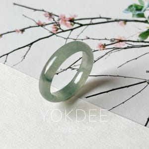 May include: A smooth, polished jade ring with a translucent pale green hue and subtle darker green inclusions. The band features a rounded profile and a glossy finish, reflecting light across its surface. The text YOKDEE JEWELLERY is visible.