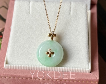 Puede incluir: Un collar de cadena de oro con un colgante circular de jade verde pálido. El colgante está adornado con un pequeño dije de oro en forma de trébol con un diamante diminuto en el centro, y un dije de trébol a juego une el colgante a la cadena.