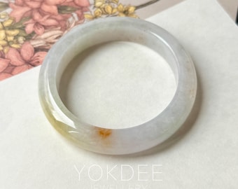 May include: A jade bangle bracelet with a translucent appearance. The bracelet features a blend of pale green, white, and a touch of orange-brown hues. The text "YOKDEE JEWELLERY" is visible.