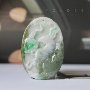 May include: Oval jade pendant with a carved design featuring a horse and cloud motifs. The pendant is primarily white with green accents, and has a hole at the top for a necklace. The text "YOKDEE JEWELLERY" is at the top.
