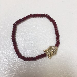 May include: A red beaded bracelet with a gold Buddha charm. The Buddha charm has a detailed face and is adorned with small clear stones.
