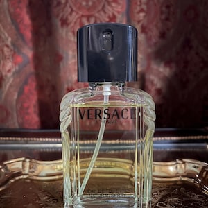 May include: A clear glass bottle of Versace L'Homme Eau de Toilette Natural Spray. The bottle has a black cap and a rectangular shape. The fragrance is a light amber color. The bottle is sitting on a gold-colored tray.