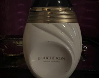 May include: A white, egg-shaped perfume bottle with a black, dome-shaped cap and a gold-toned band at the neck. The bottle features a textured, swirling design on the surface and the text BOUCHERON EAU DE PARFUM printed in gold on the front.