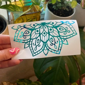 May include: A teal and blue iridescent vinyl decal of a mandala design. The decal is on a white background and is being held by a hand.