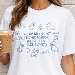 Può includere: T-shirt bianca con un design blu a tema cani. La maglietta presenta varie illustrazioni di cani e il testo "Apparently I'm Not Allowed to Adopt All the Dogs.... Rude, But Okay." La parte inferiore della maglietta ha il testo "SVG & PNG Design."
