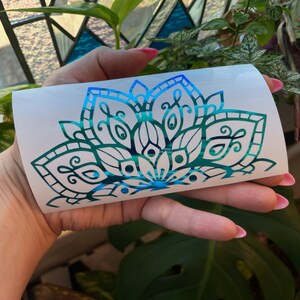 May include: A teal and blue iridescent vinyl decal of a mandala design. The decal is on a white backing.