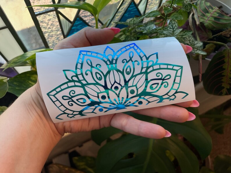 May include: A teal and blue iridescent vinyl decal of a mandala design. The decal is on a white backing.