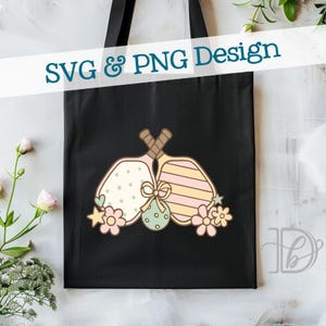 May include: Black tote bag featuring a colorful design of two pickleball paddles with decorative elements. The paddles are adorned with polka dots, stripes, and floral accents. The text "SVG & PNG Design" is displayed across the top.