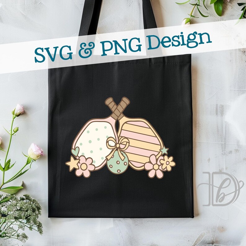 May include: Black tote bag featuring a colorful design of two pickleball paddles with decorative elements. The paddles are adorned with polka dots, stripes, and floral accents. The text "SVG & PNG Design" is displayed across the top.