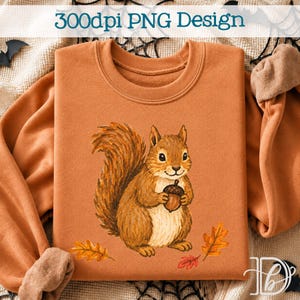 Puede incluir: Una sudadera naranja con un gráfico central de una ardilla marrón sosteniendo una bellota, rodeada de pequeñas hojas de otoño naranjas y rojas. El texto 300dpi PNG Design está impreso en la parte superior en color azul oscuro.