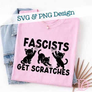 Peut inclure: Un t-shirt rose clair avec le texte "Fascistes Get Scratches" et trois illustrations de chats noirs. Les chats ont des griffures. Le t-shirt est plié sur une veste en jean.