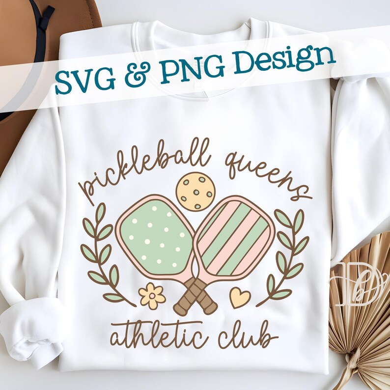 May include: White sweatshirt with a "pickleball queens athletic club" design. The design includes two pickleball paddles, a ball, and floral elements in pastel colors. The text "SVG & PNG Design" is at the top.